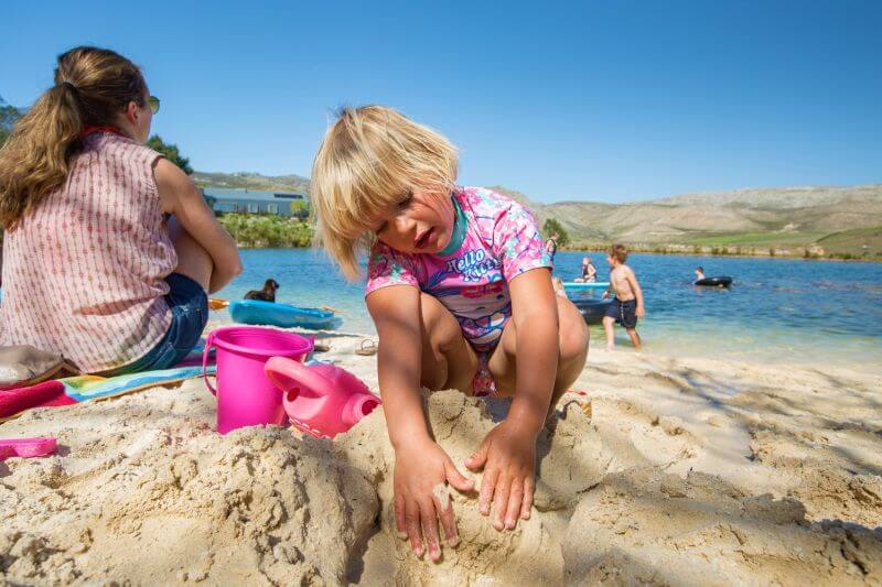 A girl building a sandcastle on the lakeside beach, others playing in the water behind, Old Mac Daddy, Elgin Valley
