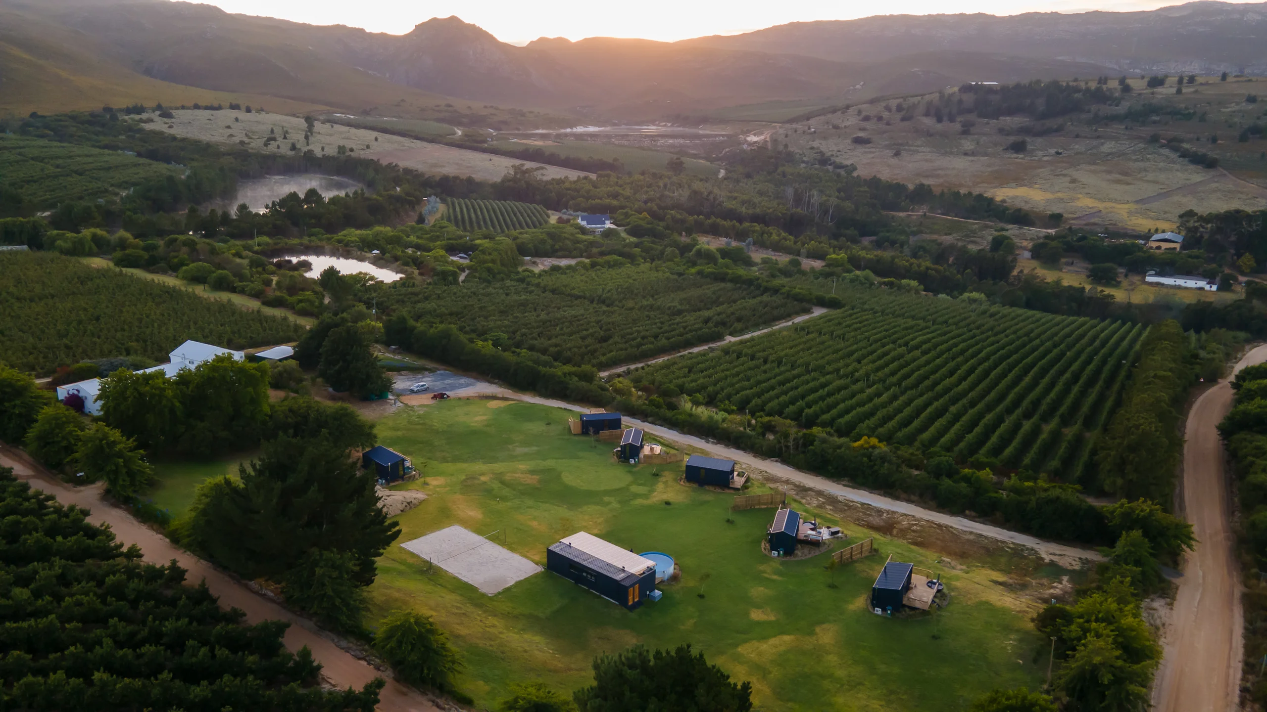 The estate from the air at golden hour: Klein Paradys cabins on the lawn, orchard rows and dams, mountains behind, Old Mac Daddy, Elgin Valley