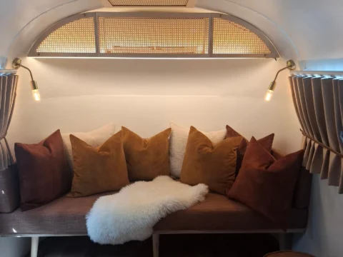 Sovereign 59: a banquette heaped with velvet cushions and a sheepskin throw under an arched cane window, inside the Airstream, Old Mac Daddy, Elgin Valley