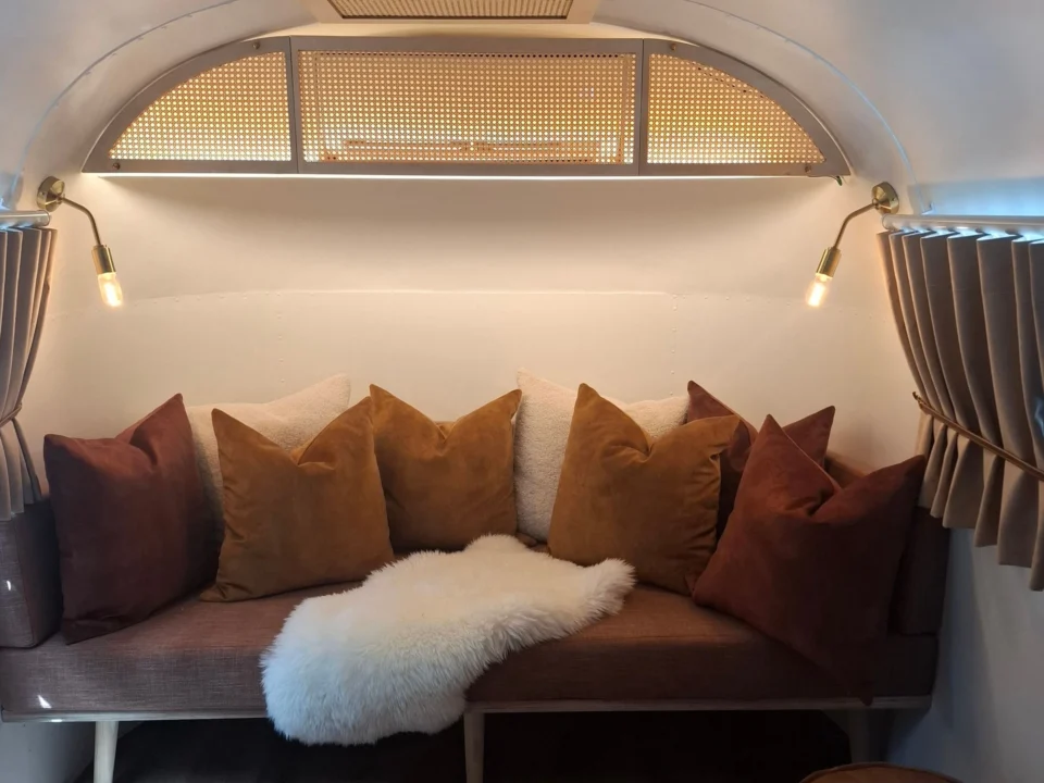 Sovereign 59: a banquette heaped with velvet cushions and a sheepskin throw under an arched cane window, inside the Airstream, Old Mac Daddy, Elgin Valley