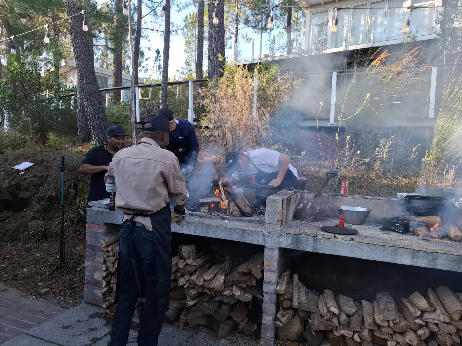 Teams cooking potjiekos over open fires at the long brick braai under the pines, firewood stacked below, Old Mac Daddy, Elgin Valley