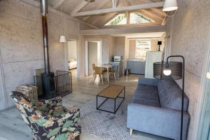 Open-plan villa living area with a wood stove, a floral armchair and a kitchen beyond, Old Mac Daddy, Elgin Valley