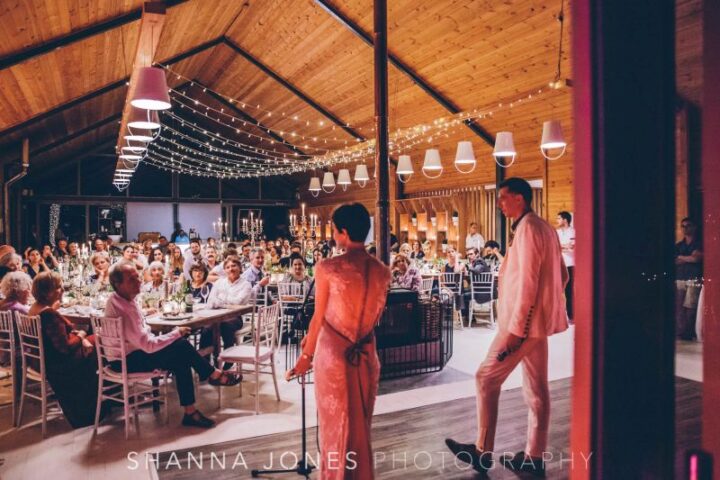 A couple speaking to guests at long tables in the Barn under festoon lights, Old Mac Daddy, Elgin Valley